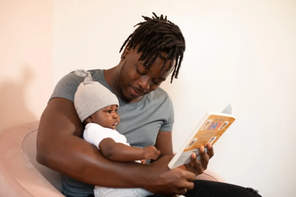 A father smiling at his baby while reading a book to the baby.