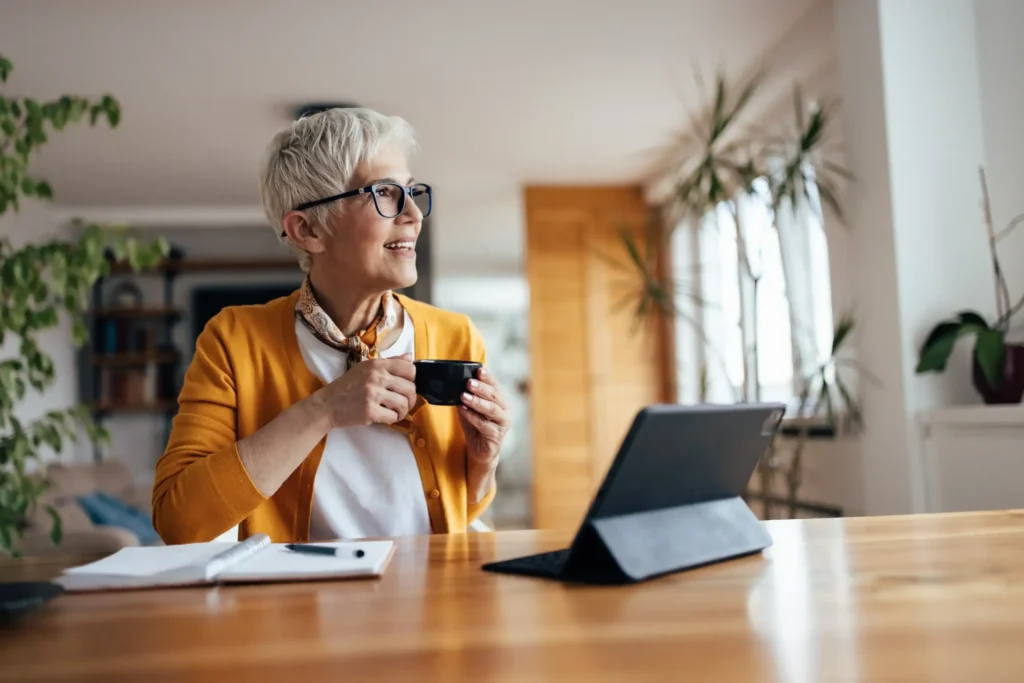 A confident older woman smiling while drinking coffee and using a tablet, representing the emotional freedom and lifestyle benefits of divorce after 50 years of marriage.